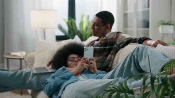 家の植物を通して見るアフリカ系アメリカ人幸せな家族カップルリラックスしていますカウチマン作業ゲームノートパソコンの女性妻と膝の上に横たわる夫スクロール携帯電話のソーシャルメディアホームソファ — ストック動画