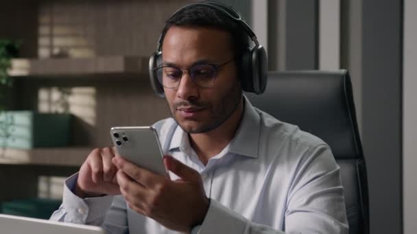 アメリカ人ビジネスマンヘッドフォンでアラビア語の労働者の上司は 携帯電話を使用して音楽を聞くオフィステーブルでサウンドアプリで曲を選択します — ストック動画