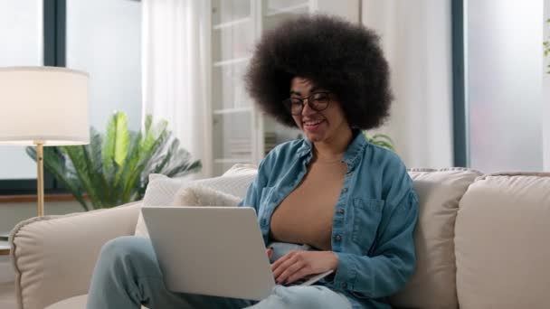笑顔アフリカ系アメリカ人のフリーランスユーザーの女性がノートパソコンのオンラインサービスを使用してソファにメッセージを入力幸せな笑顔の学生の女の子が自宅で働いているインターネットのソーシャルメディアを閲覧チャット — ストック動画