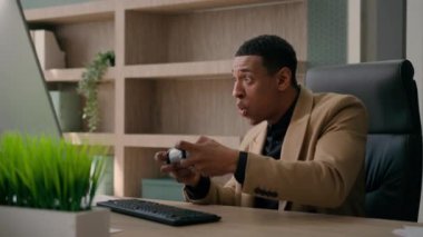 Komik ofis çalışanı mutlu Afrikalı Amerikalı erkek oyuncu kumanda kumandası bilgisayar oyunu erkek iş adamı konsol futbol video oyunu oyun bağımlılığı iş yerinde eğleniyor.