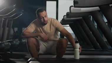 Yorgun Afrikalı Amerikalı adam koşu bandında oturur spor salonunda sıkı antrenman yaptıktan sonra rahatlar terli sporcu koşucusu su şişesi içer susuzluk giderici vücut ferahlatıcı su içer