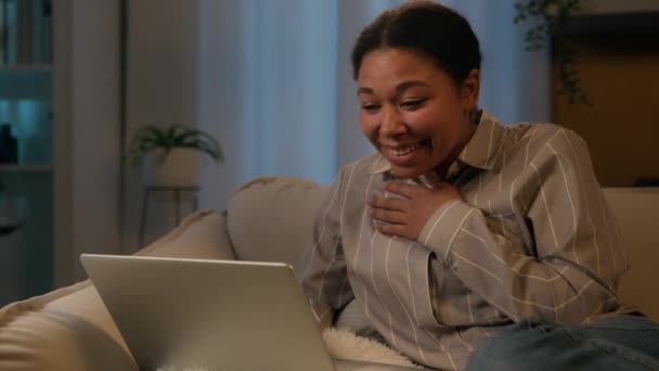 笑いアフリカ系アメリカ人女性笑顔リラックスした女の子女性の残りの部分でソファに夜の家のレジャーでノートパソコンの時計映画笑いを楽しみます素晴らしいニュースオンラインビデオ呼び出しで隔離 — ストック動画