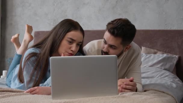 白人夫婦の住宅所有者家族思慮深い夫と妻のショッピングオンラインベッドの上で選択アパートノートパソコンアプリケーション上で疑わしい男と女の話寝室でインターネット購入を計画考えて — ストック動画