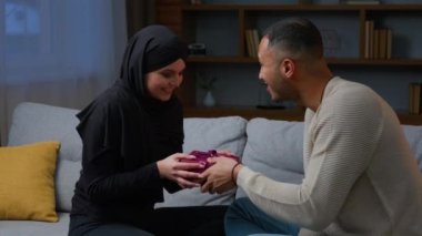 Afrikalı Amerikalı erkek, tesettürlü, heyecanlı eş kucaklayan Müslüman kadına sürpriz hediye kutusu verdi. Birbirine aşık mutlu çift evde romantik sevgililer gününü kutluyor.