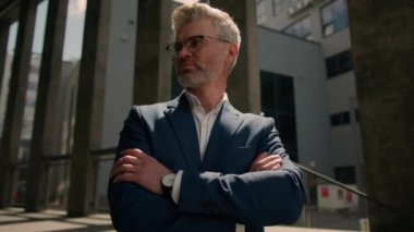 Şehrin dışında iş portresi kapatılıyor. Orta yaşlı beyaz iş adamı dişlek, kamera ofis merkezi binası önünde gülümsüyor. Erkek girişimci lideri gülümsüyor.