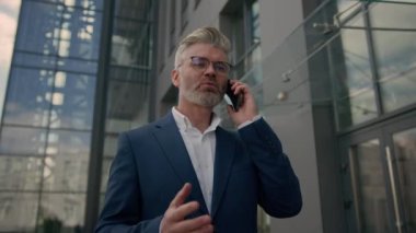 Yetişkin orta yaşlı işadamı şehirde yürüyor cep telefonuyla konuşuyor kıdemli yetişkin iş adamı yatırımcısıyla görüşüyor cep telefonu girişimcisiyle konuşuyor ofis binasının yakınında yürüyor.