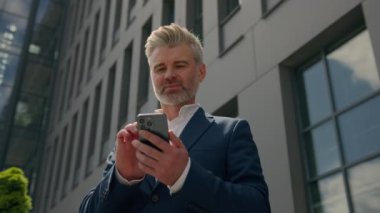 Orta yaşlı, olgun iş adamı kıdemli işadamı mutlu gülümseyen cep telefonu sohbeti uygulaması akıllı telefon tarama kripto cep telefonu sipariş taksisi ofis binası yakınlarındaki alt manzaralı.