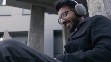 Arap adam Hintli girişimci işadamı, milenyum boyunca kulaklıkla gülümseyen öğrenci, ofis binasının yanında dizüstü bilgisayarla çalışıyor.