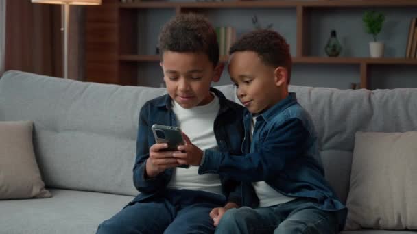 两个非洲裔美国兄弟姐妹坐在沙发上 用手机聊天 滚动智能手机 玩网络游戏 孩子们 孩子们 兄弟们一起在家里浏览手机应用程序聊天 — 图库视频影像