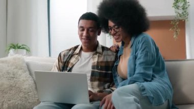 Mutlu aile çifti, Afrikalı Amerikalı koca kanepe üzerinde kadın kanepe dizüstü bilgisayarı internet alışverişi internet uygulama mağazasından tatil biletleri alarak mal siparişi teslimatını birlikte seçin.