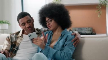 Mutlu Afro-Amerikan çift kanepesi internetten alışveriş yapan ürünleri seçiyor. İnternet mağazası uygulamalarında konuşuyorlar. Akıllı telefon siparişi, birleşik bütçe.