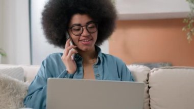 Mutlu gülümseyen Afrikalı Amerikalı serbest çalışan kadın müşteri alıcısı cep telefonu alışverişi sipariş yiyecek teslimatı laptopla çalışıyor projeyi tartışıyor evdeki koltukta akıllı telefon konuşması yapıyor.