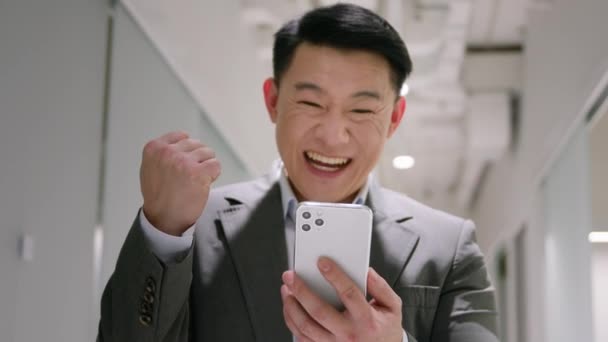 快乐惊讶的40多岁的中年男人看着办公室里的手机 男性获奖者亚洲韩国商人赢得了商业成功阅读令人惊叹的新闻是的 令人惊奇的是智能手机赢得了成就 — 图库视频影像