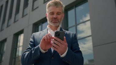 Orta yaşlı, olgun iş adamı, sohbet eden cep telefonu işvereni internet bahsini kazanma konusunda şaşkınlığa düşmüş, ofis binası yakınlarında akıllı telefon zaferi kazanmış.