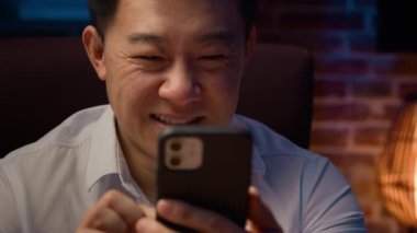 Mutlu gülümseyen Asyalı orta yaşlı iş adamı Koreli yetişkin işadamı Çinli işçi Japon işvereni gece ofiste cep telefonlarını karıştırıyor akıllı telefon konuşmaları yapıyor.