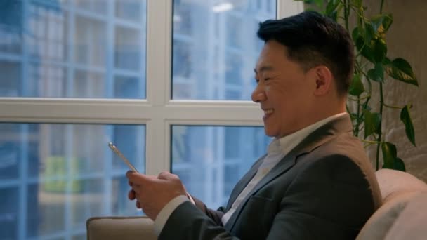 中年アジアのビジネスマン幸せな笑顔多人種の中国人男性オフィスで携帯電話でインターネットソーシャルメディアを閲覧喜びの男性起業家使用ガジェット携帯電話ビジネス地区ウィンドウビュー — ストック動画