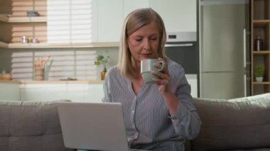 Evde dizüstü bilgisayarda yazıp kahve içen olgun beyaz bir kadın kanepe üzerinde yaşlı bir iş kadını sohbet mesajları yolluyor internetten e-posta alışverişini kontrol ediyor sosyal ağda keyif çatıyor.