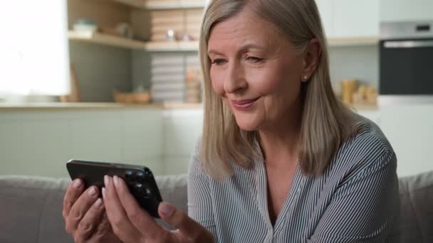 笑顔高齢者の白人女性は携帯電話の画面を見て笑ってインターネット上で面白いビデオを見て写真を閲覧楽しいオンラインを持っていますホームソファでスマートフォンを使用してソーシャルネットワーク上で良いニュースを読んで — ストック動画