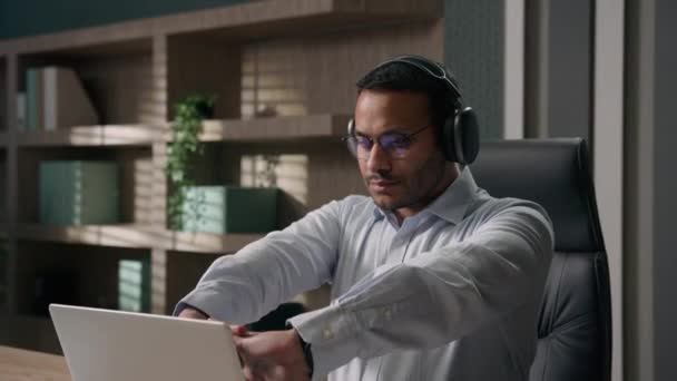 インドのビジネスマンストレッチ手の指は仕事のラップトップを開始しますアラビア人男性アメリカ人のオフィスワーカービジネス雇用主入力ワーキングコンピュータは ヘッドフォンで音楽を聞くやる気ムードソングオーディオサウンド — ストック動画