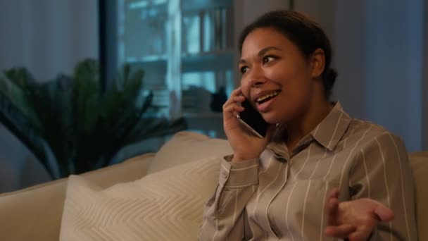 笑顔アフリカ系アメリカ人女性トーク携帯電話注文食品配達良いニュースを聞きます感情的な話オンソファ励起女性ビジネス女性気楽なビジネス会話に夜の家 — ストック動画