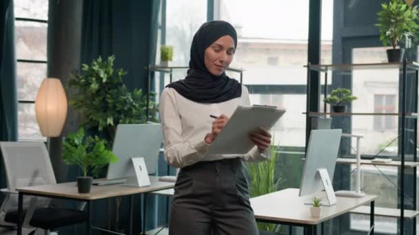伊斯兰穆斯林印度女商人商业女经理商业招聘女孩戴头巾在办公室里检查写在笔记本上的字条签署文件保险清单看着相机微笑 — 图库视频影像