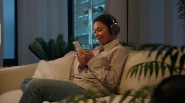 Akşamları kanepede kulaklık takmış gülümseyen Afrikalı Amerikalı kadın modern teknoloji kadın müzik aşığıyla evde şarkılar dinliyor cep telefonlu kız dinleniyor oturma odasında kanepede dinleniyor.
