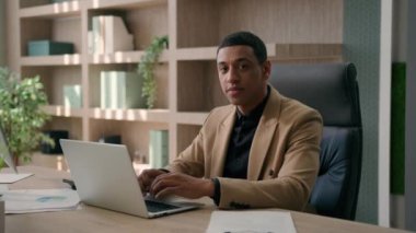 Gülümseyen iş adamı Afrikalı Amerikalı erkek girişimci iş adamı yazılım mühendisi ofis işyerindeki dizüstü bilgisayarında proje yazıyor mutlu etnik adam yüzünü kameraya çevir dişlek gülümsemeye bak