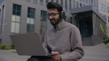 Arap Hindistanlı işadamı, şehir daktilo şirketinde dizüstü bilgisayarda çalışan iş adamı, açık havada gülümseyen adam, kulaklıkla müzik dinleyen, ofis binası yakınlarında şarkı söyleyen adam.