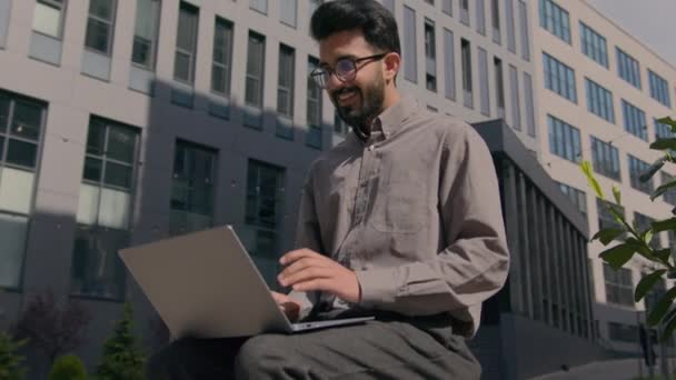 幸せなインドのビジネスマンは ラップトップ屋外都市の仕事で働くオフィスビルの外で男性フリーランスのビジネス雇用者を驚かせた興奮したアラブ人男性はオンライン勝利の達成を祝う — ストック動画