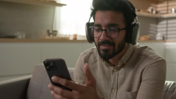 无忧无虑的印第安人房主头戴耳机在家里厨房里听音乐在耳机里听音乐和手机一起欣赏音乐放松有趣的家伙放松唱歌听音乐在线音频服务 — 图库视频影像