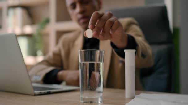 過労した病気アフリカ系アメリカ人のビジネスマンは 水のガラスの中に鎮痛剤の錠剤を投げるオフィステーブルの上に飲み物の泡が付いている固定薬を適用します頭痛ハングオーバー病気のビジネスマンは救済策を準備 — ストック動画