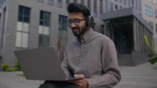 アラビア語インドの実業家の雇用主の男性起業家都市の入力ビジネスプロジェクトの仕事のラップトップで働く屋外の笑顔の男はヘッドフォンで音楽を聴くオフィスビルの近くの歌を歌うオーディオを楽しむ — ストック動画