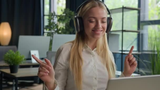 白人女性の女の子は オフィスで休憩を取りますリラックスしたビジネス女性の笑顔は ノートパソコン上のヘッドフォンで音楽を聞きますオンライン音楽サービスマネージャーは イヤホンで聞いてメロディーダンスの動きをお楽しみください — ストック動画