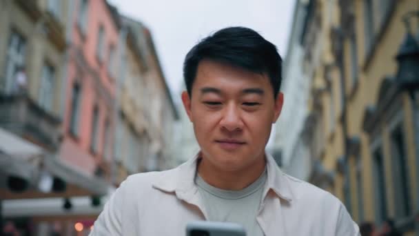 近くで笑っているアジア人男性が路上で電子メールをチェック携帯電話を使用してソーシャルメディアを閲覧幸せな韓国人男性ビジネスマンは スマートフォンでオンラインチャットメッセージを送信市内の屋外で歩く — ストック動画