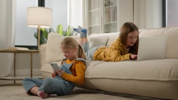 子供と近代的な技術2つの幸せな子供の女の子の姉妹の友人は自宅でモバイルやコンピュータアプリケーションを使用して遊んで楽しんで居心地の良いリビングルームでノートパソコンの携帯電話で一緒にビデオオンラインゲームをプレイ — ストック動画