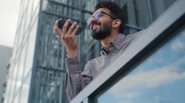 Mutlu gülümseyen Hintli işadamı işvereni telefonda sesli mesaj kaydediyor. Sesli mesajı mikrofona kaydetmek için yapay zeka cep telefonu yardımı kullanıyor. Şehirdeki hoparlörlerde akıllı telefonlar konuşuyorlar.