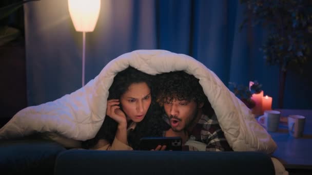 一对印度男人和阿拉伯西班牙女人在床上约会 在毛毯下的男朋友女朋友震惊地看着恐怖电影害怕害怕用手机藏着吓人的视频 — 图库视频影像