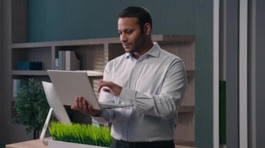 Ofisteki iş portresi yetişkin Hintli adam etnik iş adamı Arap işadamı dizüstü bilgisayarlı işveren kameraya gülümseyen online yakın bilgisayardaki işi bitirdi