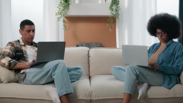 アフリカ系アメリカ人フリーランスの家族カップル作業とともに2台のコンピュータノートパソコンでホームカウチ女性男使用技術再生ビデオゲームガジェットインターネット中毒配偶者仕事遠隔でソファ — ストック動画