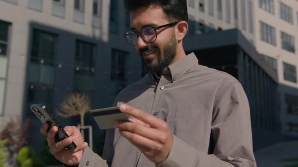 幸せなインドのアラビア人の男性は 市内の携帯電話やクレジットカードを保持屋外でお金を支払うオンラインバンキングは 興奮したビジネスマンを祝うビジネスの勝利をオンラインショッピングの成果 — ストック動画