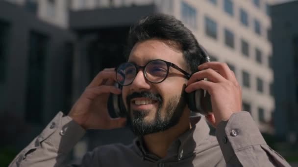 幸せな屈託のないアラビア人の男は ヘッドフォンを着用し 日当たりの良い都市の屋外で大声で音楽を聞くスマートフォンで高音をお楽しみくださいオーディオサービスをオンラインで聞いているインドのビジネスマン外のオフィス会社 — ストック動画