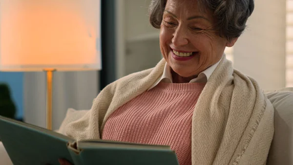 Happy smiling laughing Caucasian old woman reading bestseller book peace at home covered with blanket senior mature lady smile elderly female retired grandmother enjoy read funny novel literature