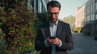 Beyaz adam şehir caddesinde yürüyor ofis binalarının yakınına gidiyor mutlu işadamı işvereni cep telefonuna mesaj atıyor medya konuşmaları yapıyor gülümseyen adam açık havada akıllı telefon kullanıyor.
