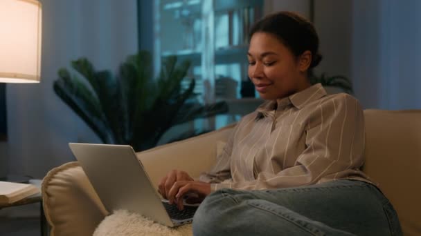非洲裔美国自由职业者女孩晚上在笔记本电脑上工作 晚上在家上网学习 在沙发上接受远距离教育 隔离微笑着在电脑社交媒体上看电影的女性 — 图库视频影像