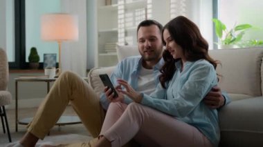 Millennial couple Caucasian man woman married husband wife at home talking using smartphone together happy family discuss offers choosing shopping online with mobile phone boyfriend showing thumb up