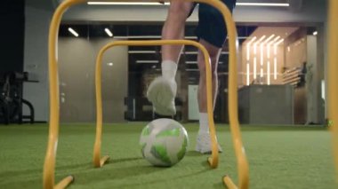 Unrecognizable man male footballer soccer player sportsman legs hit ball through arches jump over barriers training in gym playing football game on field prepare for match workout with sport equipment