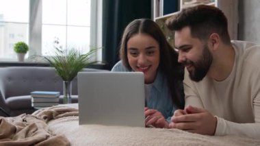 Relaxed family couple in love lying on bed with laptop shopping online choose delivery store Caucasian woman man buying internet goods booking tickets together carefree wife husband talking in bedroom