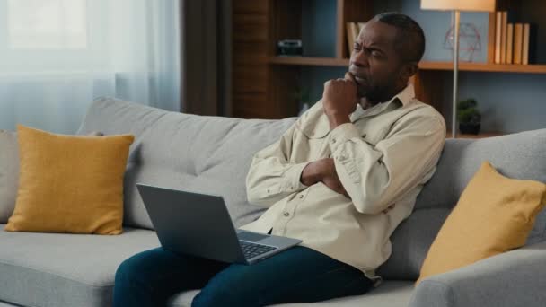 アフリカ系アメリカ人ビジネスマンフリーランサーが自宅のソファーでラップトップに取り組んで ビジネス上の問題を解決することを考える真剣なペンギン男性の新しいアイデア思考のためのインスピレーションを探す — ストック動画
