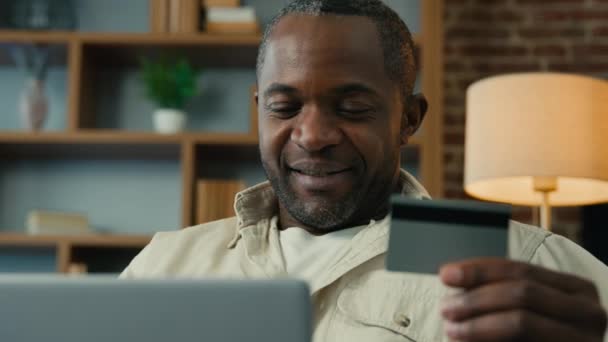 ハッピーアフリカ系アメリカ人男性は 電子バンキングアプリケーションを支払うサービスのためのオンラインストアで笑顔の男性顧客の支払いを作るリモートでラップトップで即座に簡単な支払いを使用してクレジットカードを保持しています — ストック動画