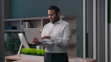 Şirket işyerinde çalışan Amerikalı bir erkek ofis çalışanı bilgisayarcı ile çalışıyor müşterilerle sohbet ediyor kameraya gülümseyen bir dizüstü bilgisayar tutuyor.
